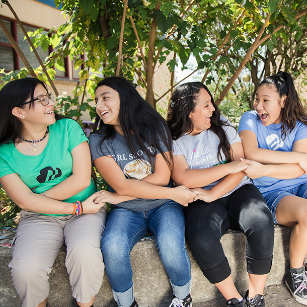 Four girls sitting on a short concrete wall. Their hands are crossed in the Girl Scout friendship circle pose.