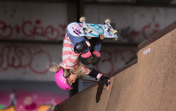 Girl riding a skateboard wearing a helmet and protetive pads. She is doing an invert on a halfpipe.