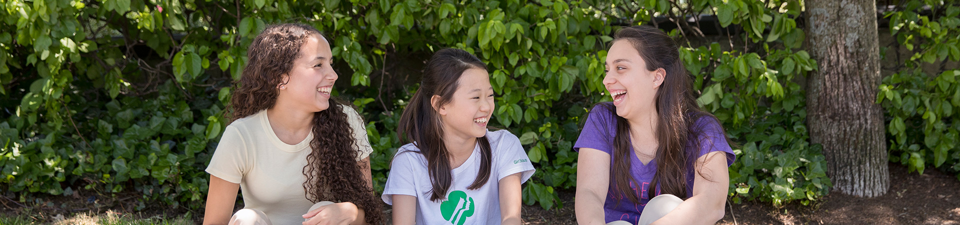  Three teenage Girl Scouts sitting outdoors in front of a greenery wall. Two are looking at the third girl, and they are all laughing. 