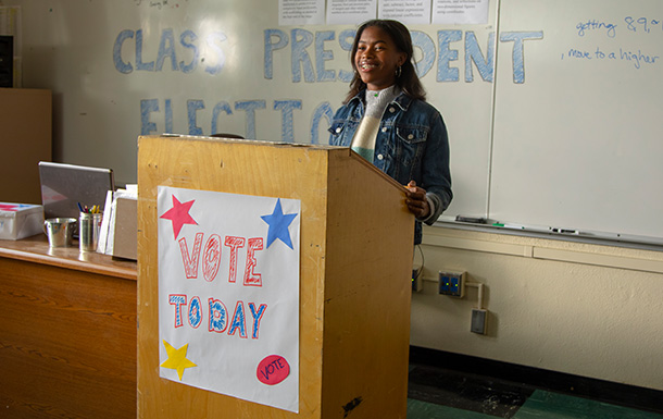 Teenage girl at a podium in a classroom with vote today poster on the podium and class president election written on whiteboard behind her.